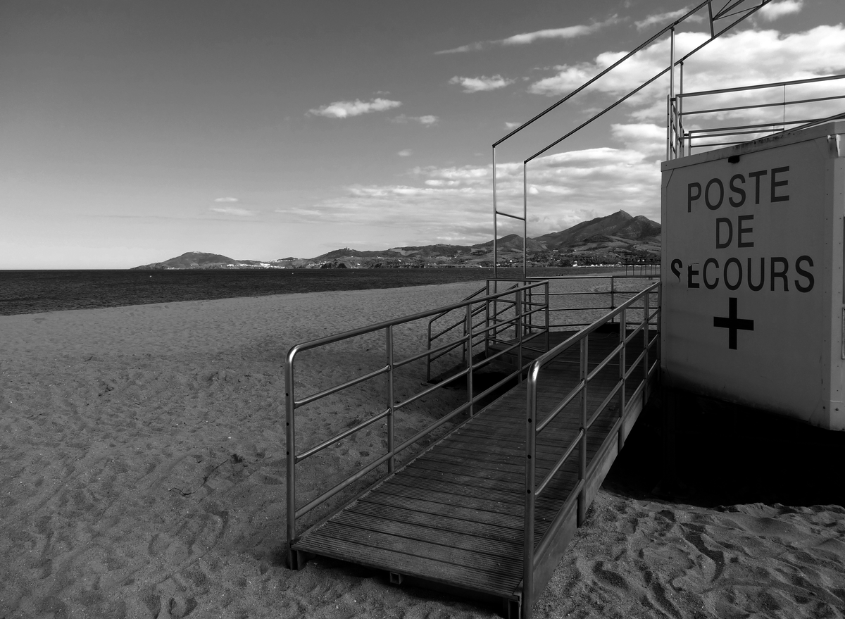 Une plage déserte, au premier plan une rampe menant à un poste de secours abandonné. Au fond, la mer et les montagnes.